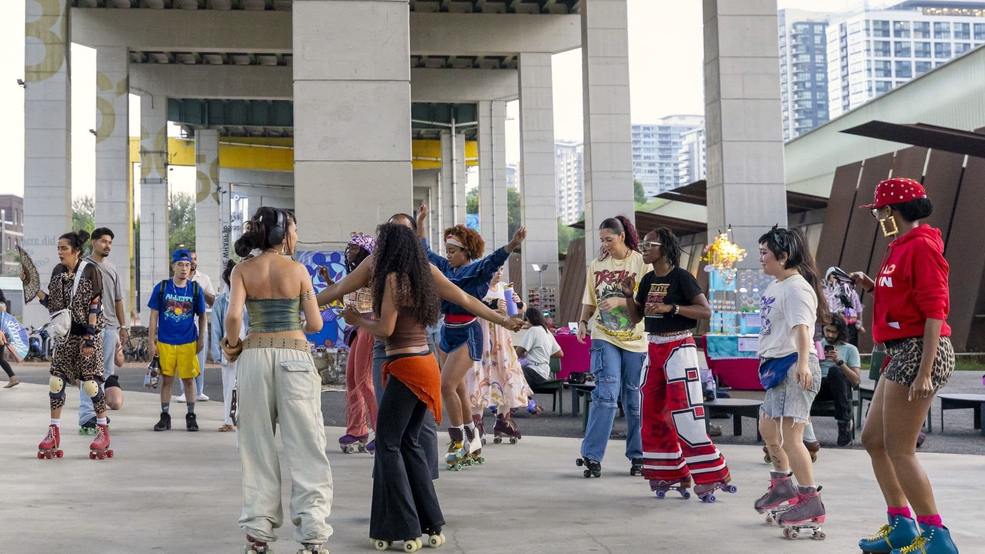 People roller skating and socializing under a highway overpass, with a lively atmosphere and urban backdrop.